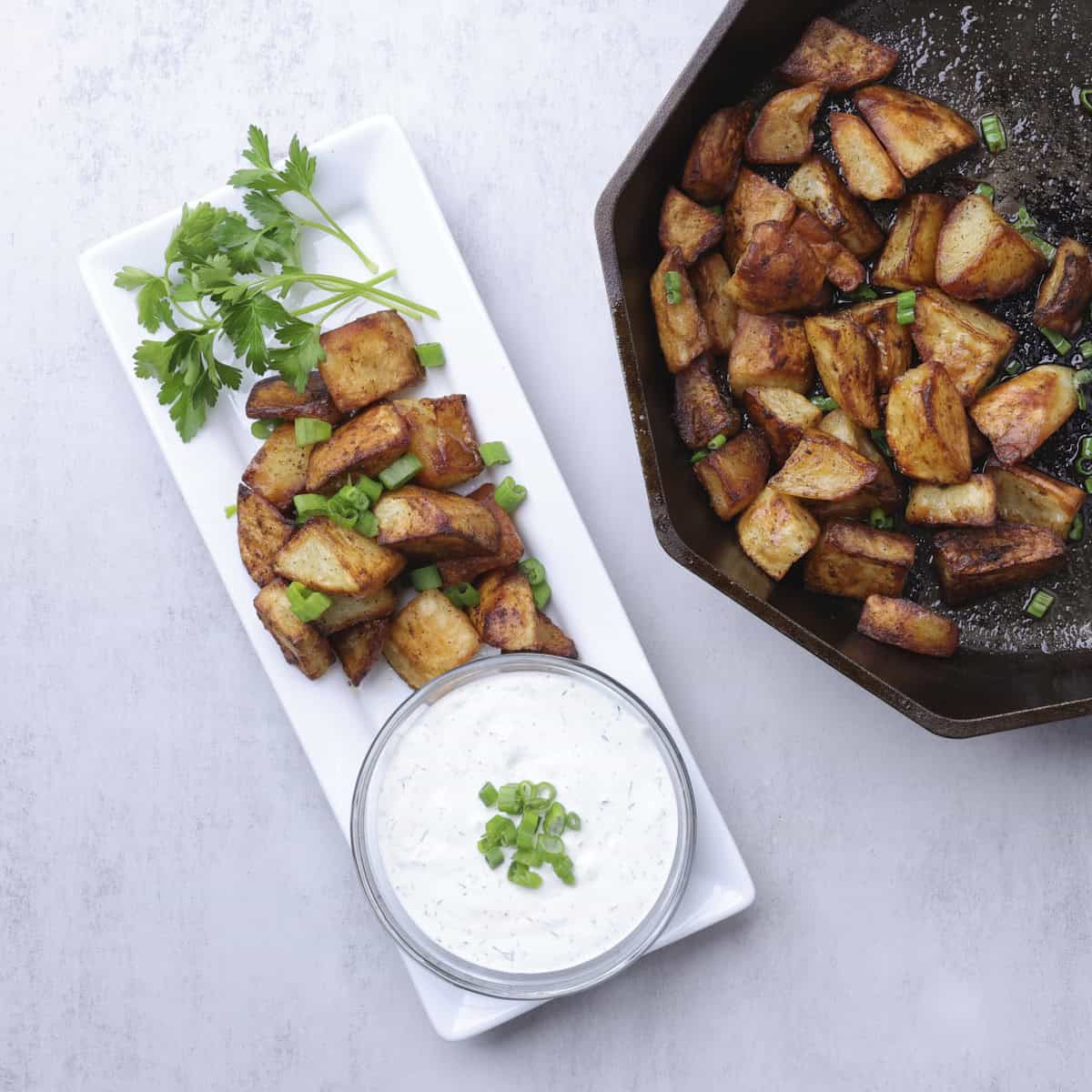 Baked Home Fries in a skillet and displayed on a white rectangular plate with a bowl of dill dip and garnished with greens.