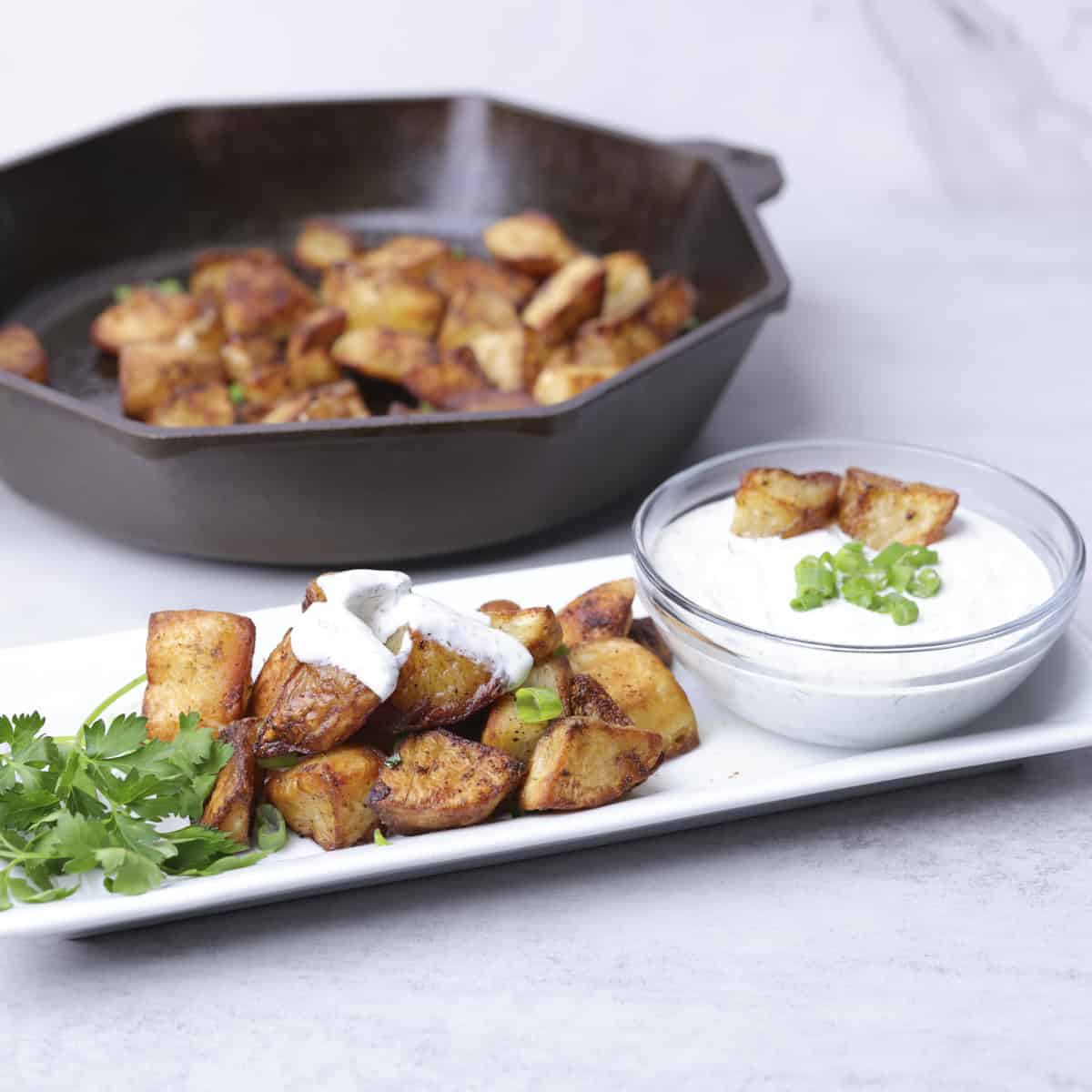 A white rectangular plate with Baked Home Fries garnished with greens, dipped in dill sauce and served with a bowl of dill sauce. A skillet of more fries in the background.