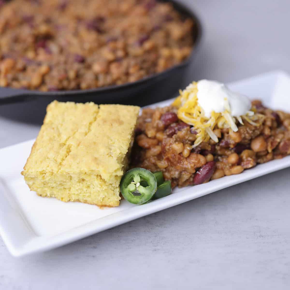 A white rectangular plate with a serving of the Baked Beans with Hamburger and Bacon, topped with cheese and sour cream and displayed with corn bread and jalapeno slices with a skillet of beans in the background.