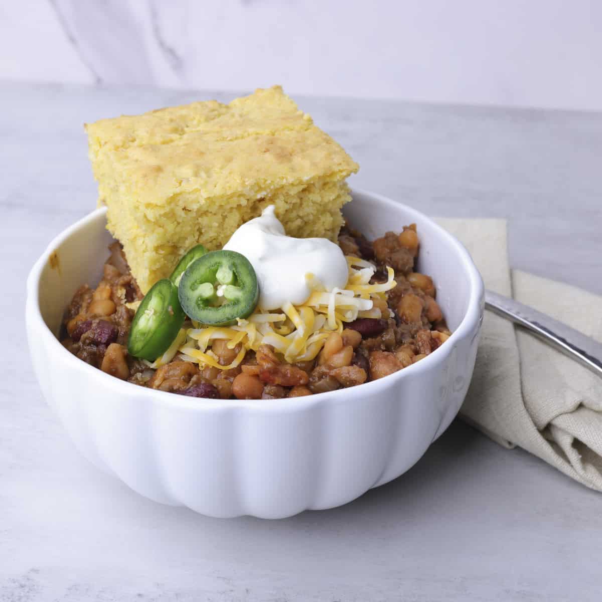 A bowl of Baked Beans with Hamburger and Bacon topped with corn bread, cheese, sour cream and jalapeno slices.