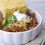 A white bowl filled with Baked Beans with Hamburger and Bacon, topped with shredded cheese, sour cream and jalapeno slices with a slice of corn bread resting on top.