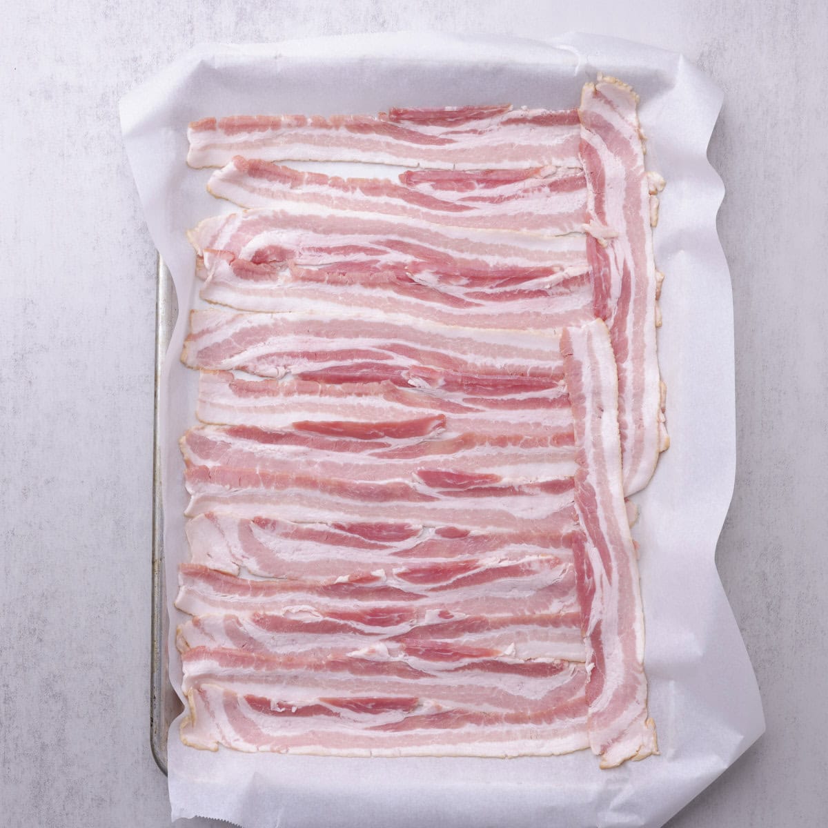A cookie sheet with a pound of bacon spread out on parchment paper, ready to bake.