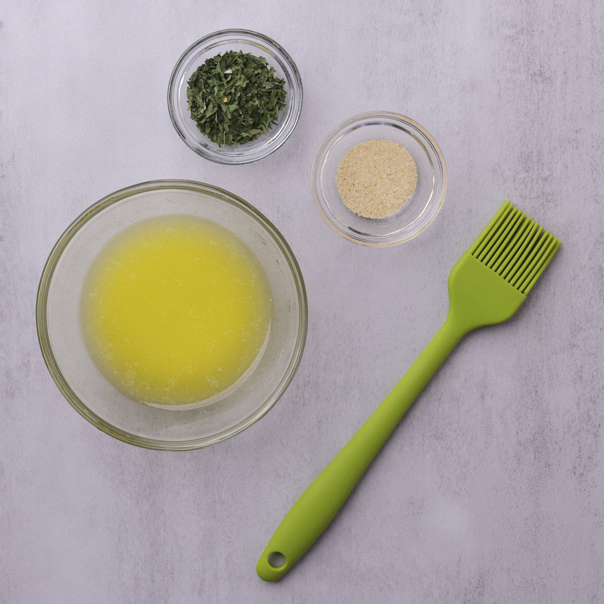 Melted butter, dried parsley and garlic powder each in their own glass bowls with a green pastry brush.