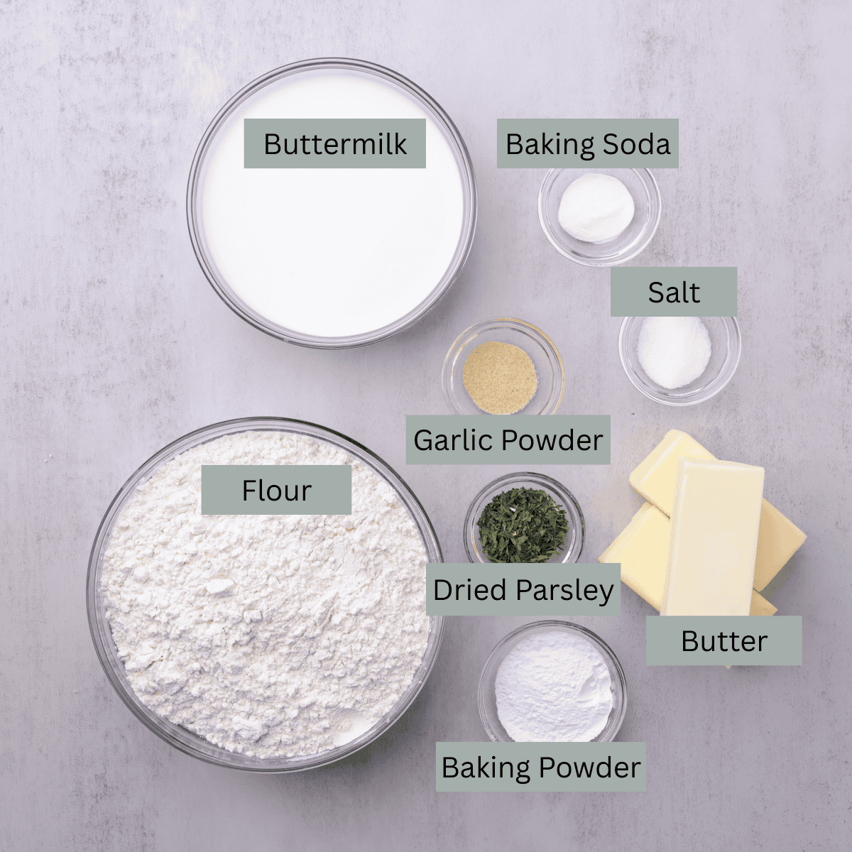 A visual display of the ingredients needed for the skillet biscuits - flour, buttermilk, baking powder, baking soda, salt, garlic powder, dried parsley and butter, each in their own bowl.