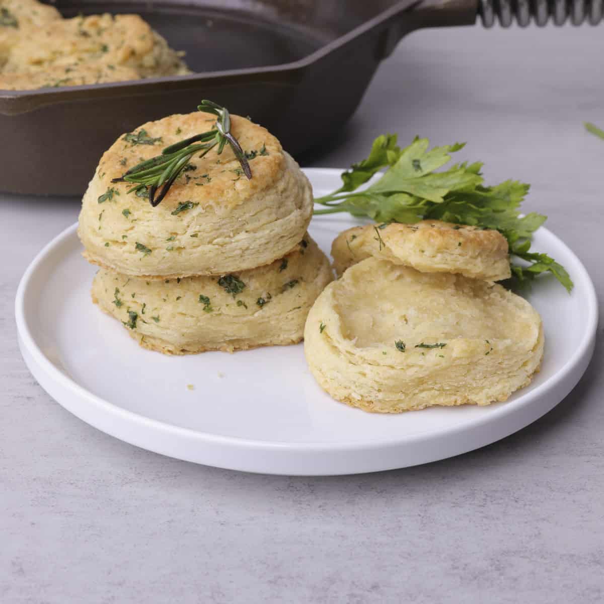 A white plate with two skillet biscuits stacked and one biscuits open faced, garnished with parsley and rosemary.