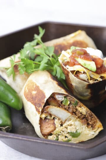 A skillet with one whole Breakfast Chorizo Burrito and one burrito cut in half and garnished with salsa, sour cream, avocado and parsley and displayed with jalapenos.