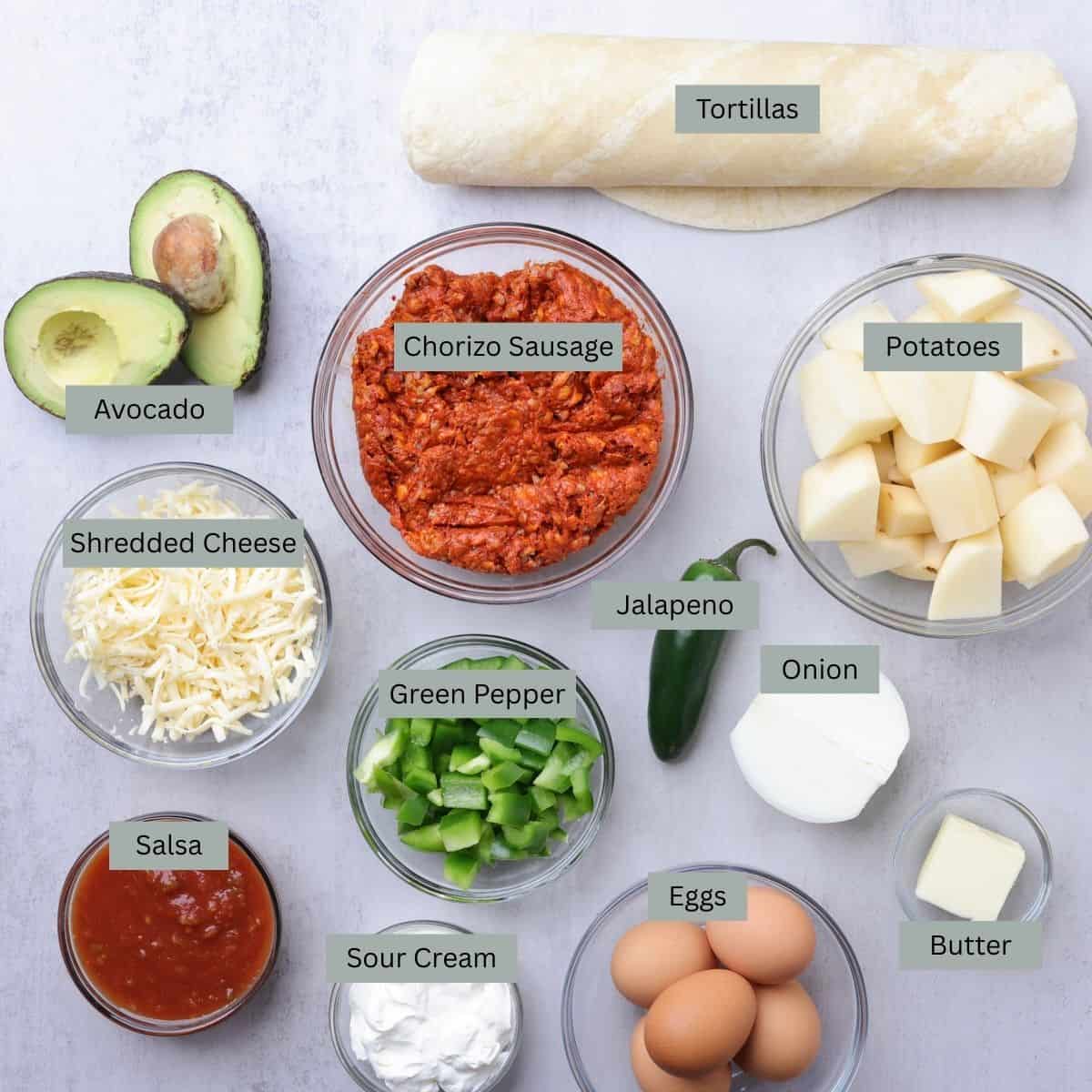 A visual display of the ingredients needed for the Breakfast Chorizo Burrito - tortillas, avocado, chorizo sausage, potatoes, cheese, bell peppers, salsa, sour cream, eggs, jalapeno, onion and butter - each displayed in their own bowl.