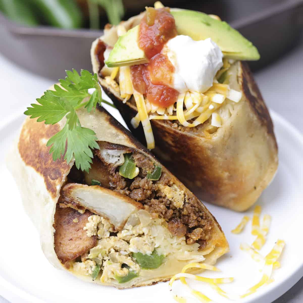 A Breakfast Chorizo Burrito Cut in half and displayed on a white plate with shredded cheese, sour cream, salsa and avocado and garnished with parsley.