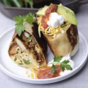 A Breakfast Chorizo Burrito Cut in half and displayed on a white plate with shredded cheese, sour cream, salsa and avocado and garnished with parsley.