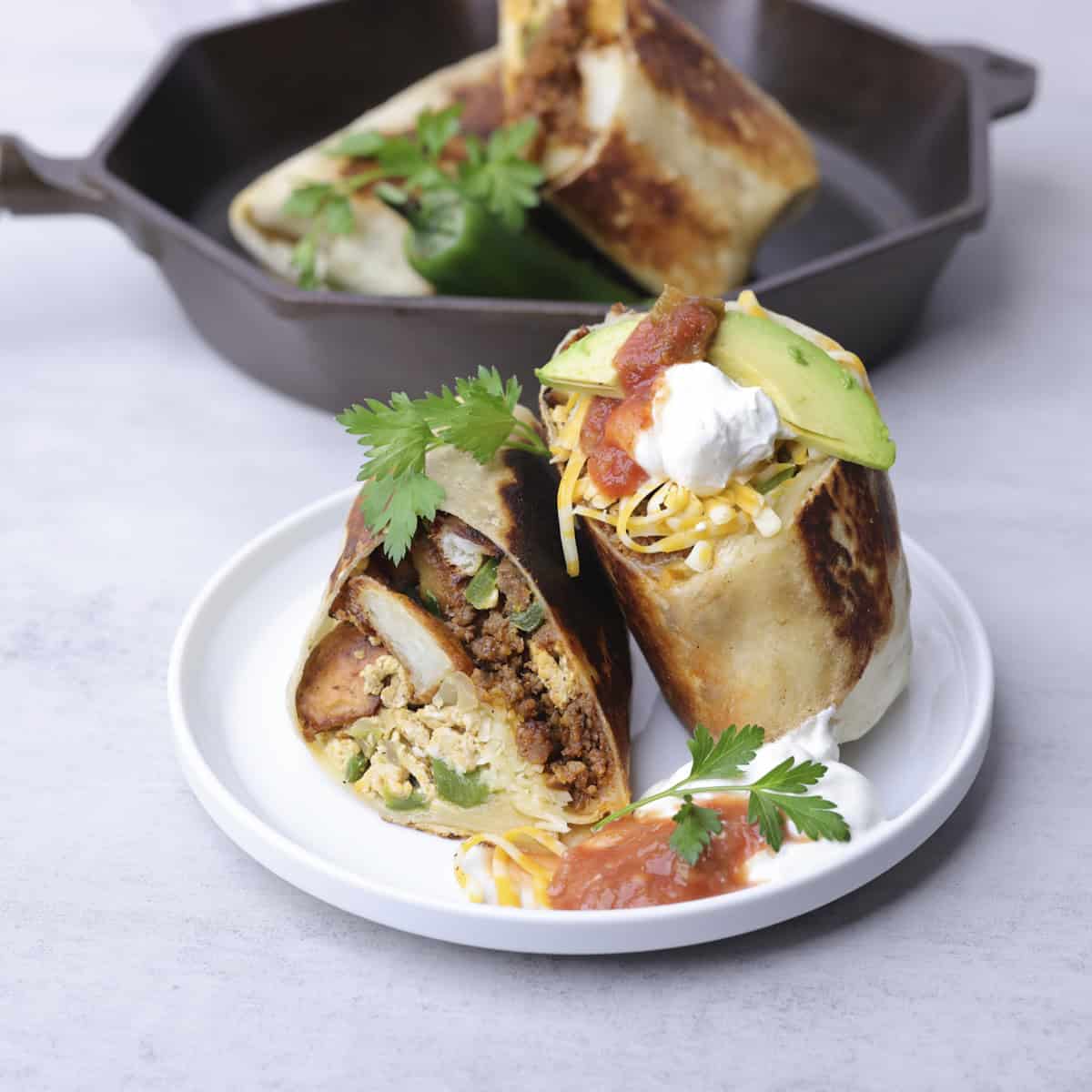A Breakfast Chorizo Burrito on a white plate topped with sour cream, salsa and avocado and garnished with parsley. Another burrito is in a skillet in the background.