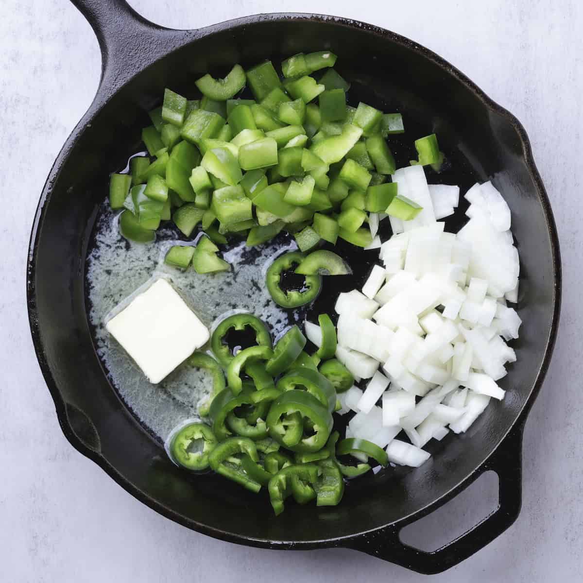 A skillet with a piece of butter melting and a pile of chopped green peppers, chopped jalapenos and chopped onion.