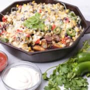 A skillet with the Chorizo Hash in it, with a portion removed displaying the potatoes, sausage and veggies in it. It is displayed with greens, salsa and sour cream.