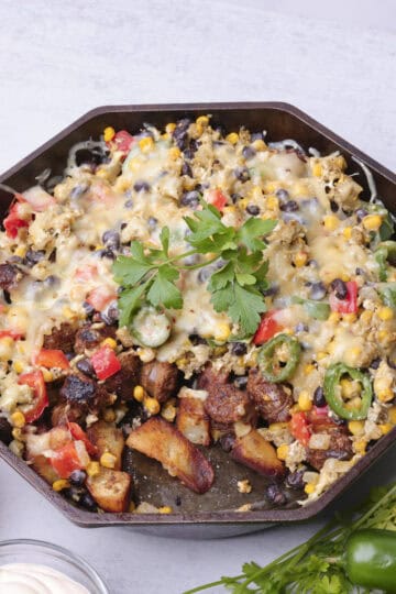 A skillet with the Chorizo Hash in it, with a portion removed displaying the potatoes, sausage and veggies in it. It is displayed with greens, salsa and sour cream.