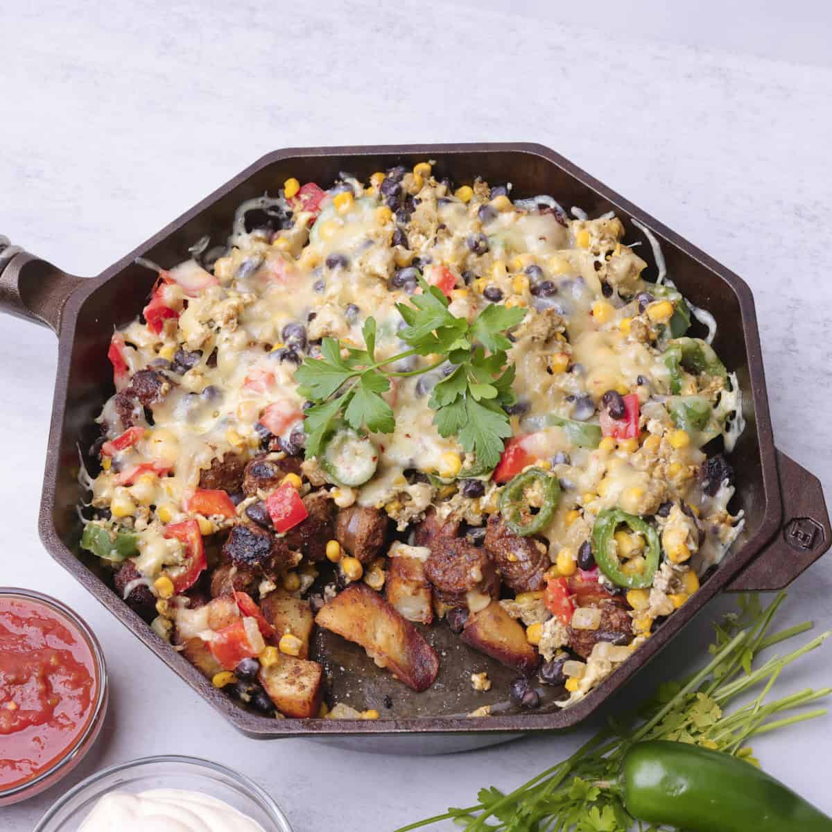 A skillet with the Chorizo Hash in it, with a portion removed displaying the potatoes, sausage and veggies in it. It is displayed with greens, salsa and sour cream.