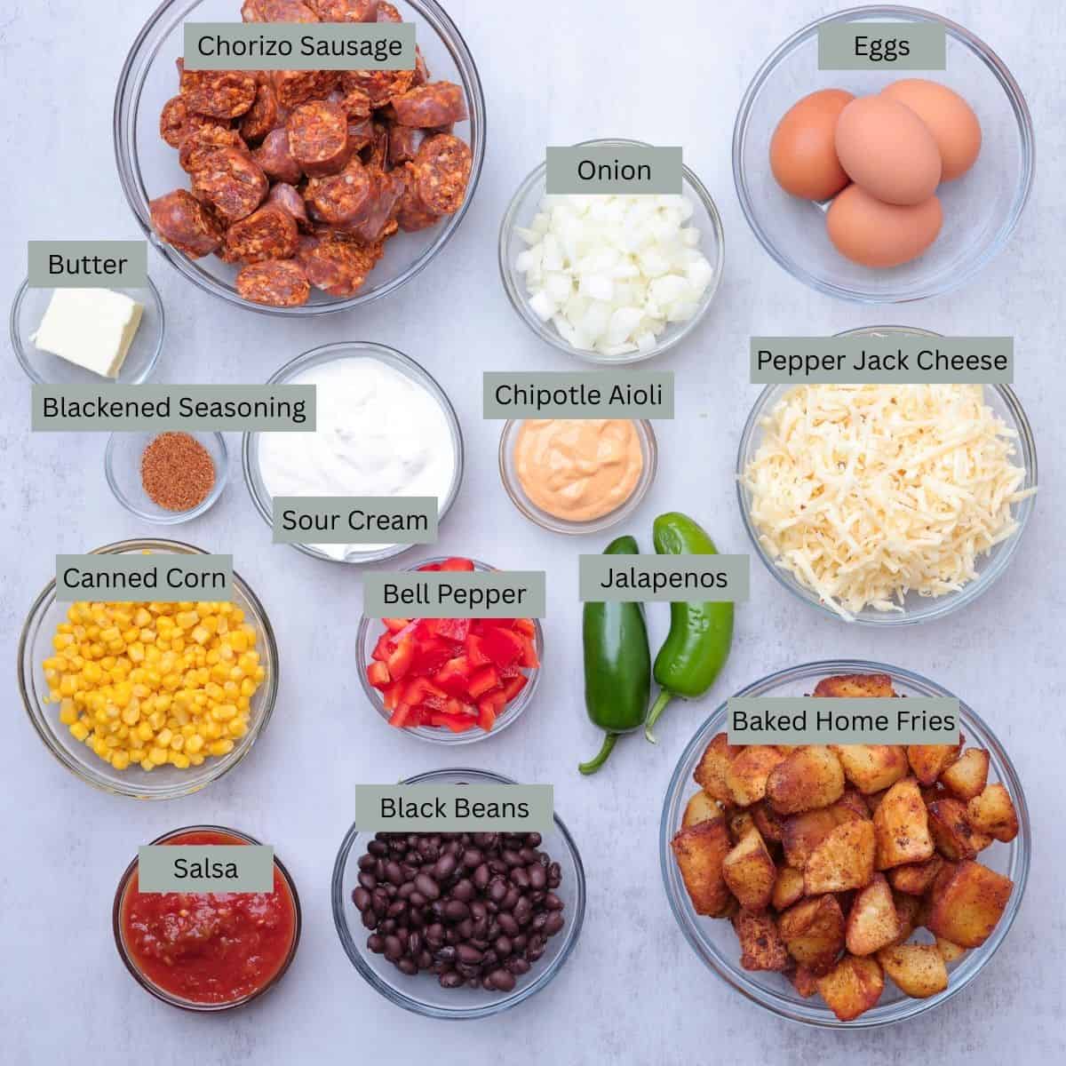 A visual display of the ingredients needed for the Chorizo Hash - Chorizo sausage, butter, onion, eggs, shredded cheese, home fries, jalapenos, black beans, bell pepper, corn, blackened seasoning, sour cream, chipotle aioli and salsa - each in their own bowl.
