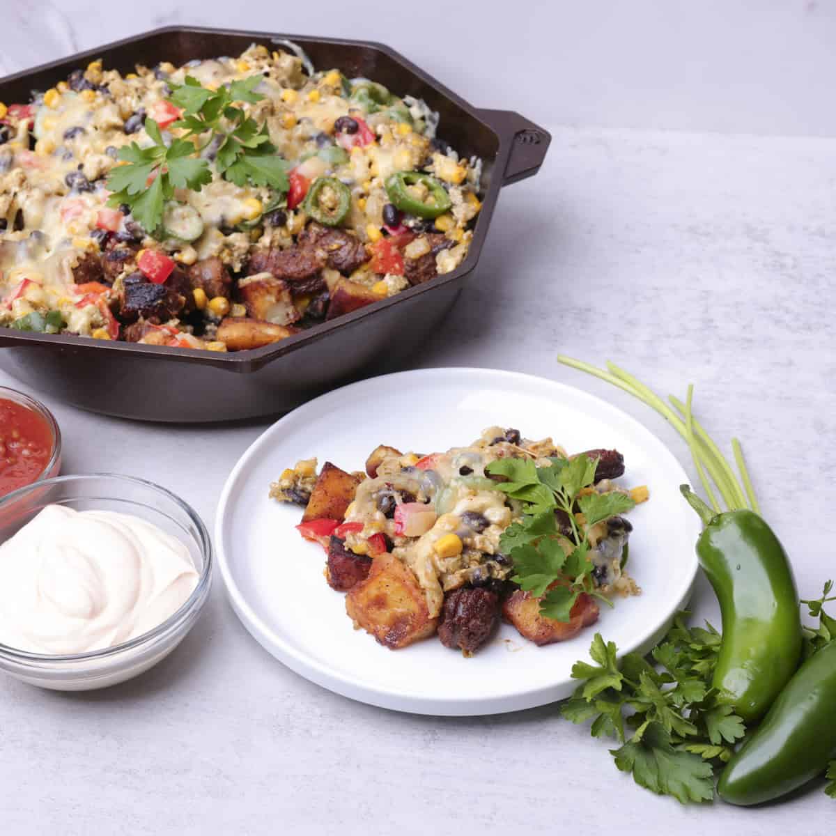 A white plate with a serving of the hash on it with a skillet containing the reaming hash in it with greens and bowls of sour cream and salsa on the side.