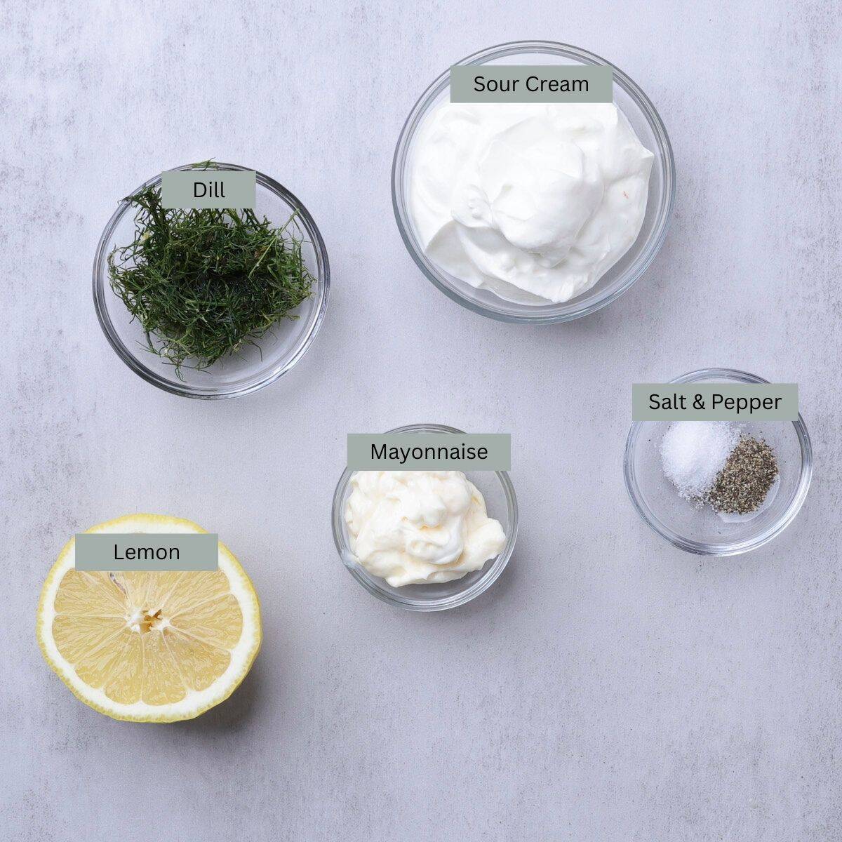 A visual display of the ingredients needed for Dill Dip - sour cream, dill, mayonnaise, salt, pepper and half a lemon - each in their own bowl.