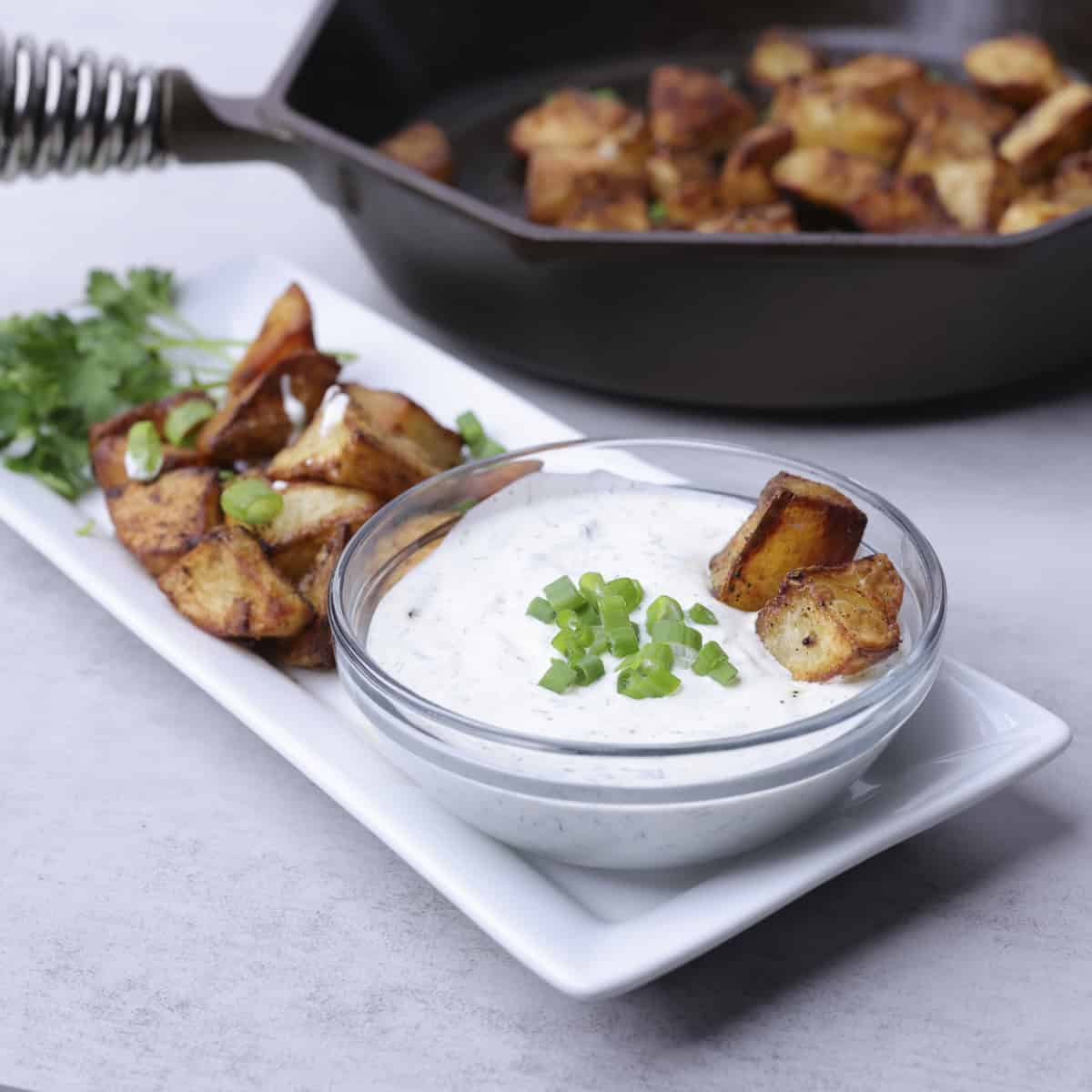 A rectangular plate with baked home fries and a bowl of dill dip on it, garnished with greens and a skillet of home fries in the background.