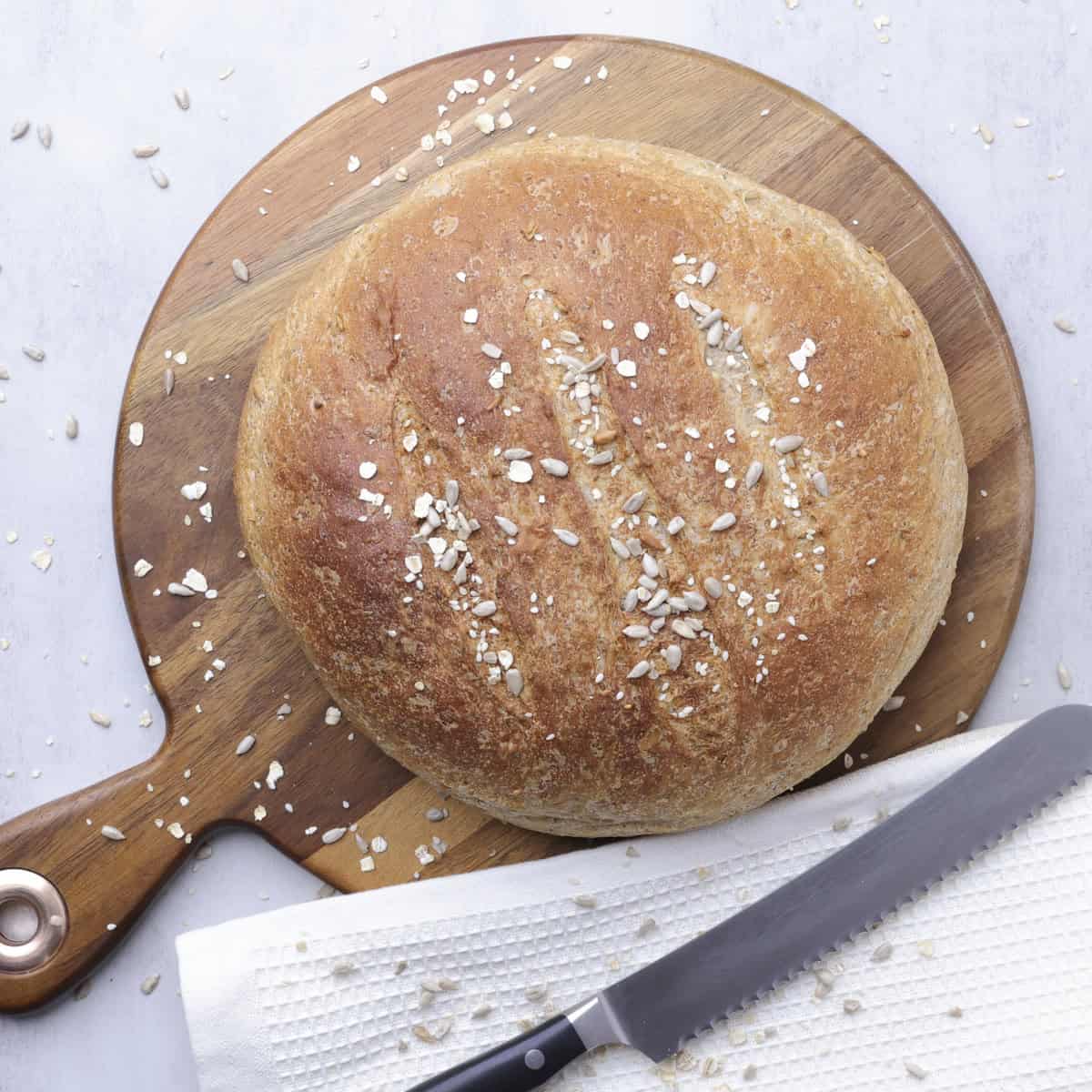 An artisan style loaf of Seeded Bread displayed on a wooden board with a tea towel and knife.