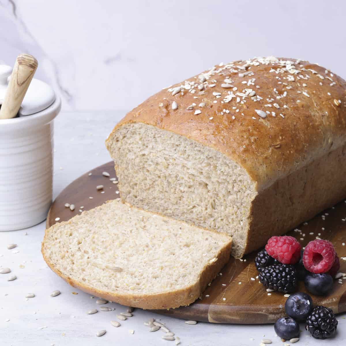 A sandwich loaf of Seeded Bread sliced and displayed on a wooden board with berries and a jar of honey.