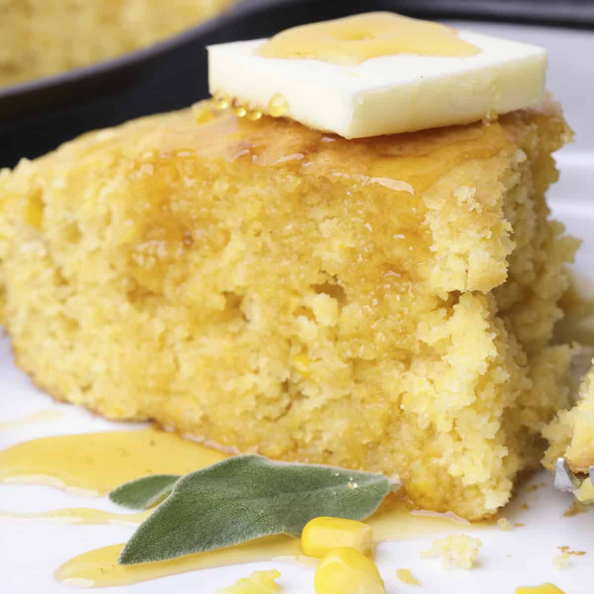 A slice of Corn Bread with a piece of butter on top, drizzled in syrup with sage leaves on the side.