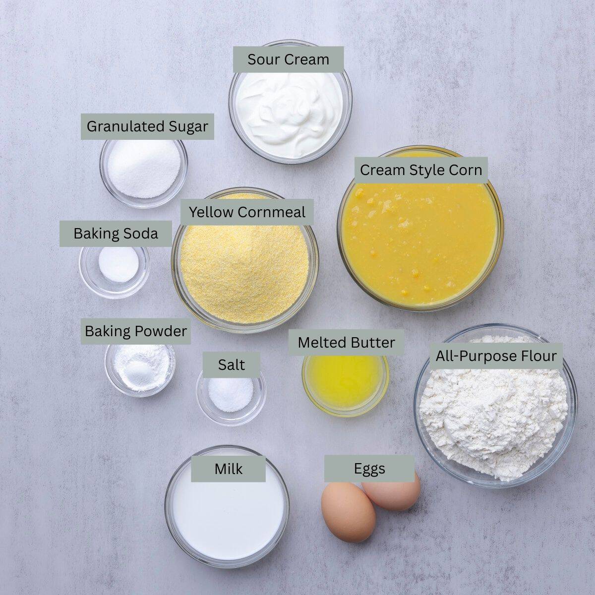 A visual display of the ingredients needed for Corn Bread with Creamed Corn - sour cream, cream style corn, flour, butter, cornmeal, eggs, milk, salt, baking powder, baking soda, and sugar - each displayed in their own bowl.