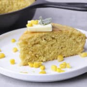 A slice of Corn Bread with Creamed Corn plated with kernels of corn sprinkled around it, topped with a slice of butter and sage leaves. A skillet of corn bread in the background.