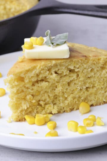 A slice of Corn Bread with Creamed Corn plated with kernels of corn sprinkled around it, topped with a slice of butter and sage leaves. A skillet of corn bread in the background.