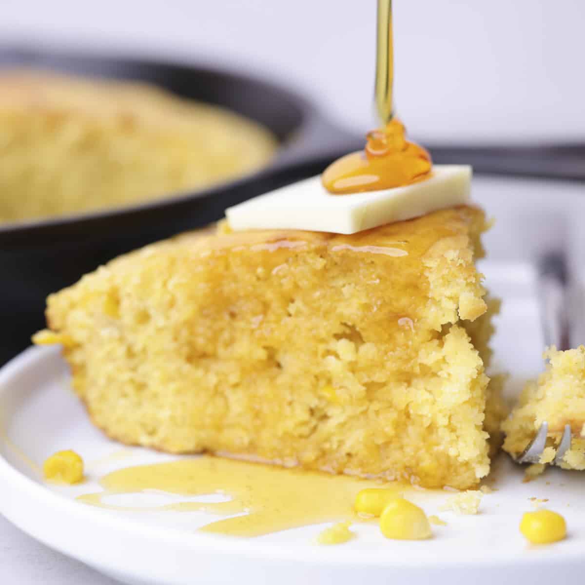 A plated piece of corn bread topped with butter with syrup being poured over it.