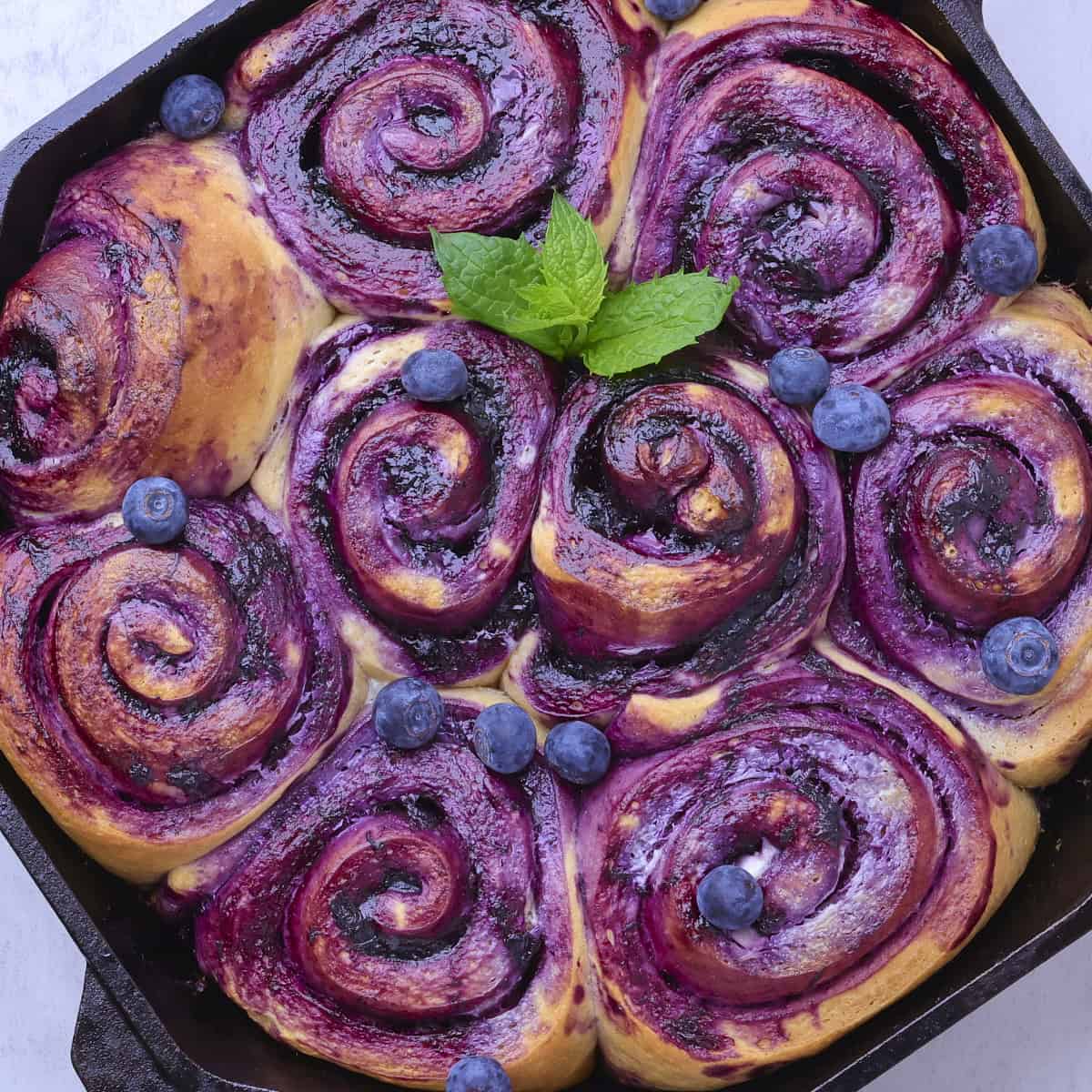 A skillet with blueberry cinnamon rolls baked to golden brown, garnished with mint leaves and sprinkled with fresh blueberries.