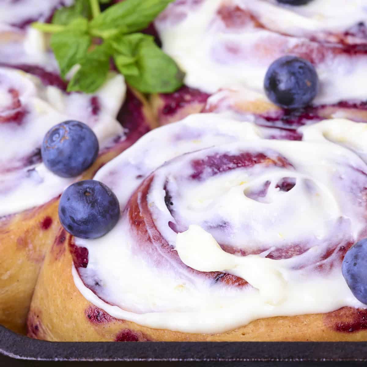 A close up shot of the Blueberry Cinnamon Rolls, topped with lemon cream cheese frosting and garnished with fresh blueberries and greens.
