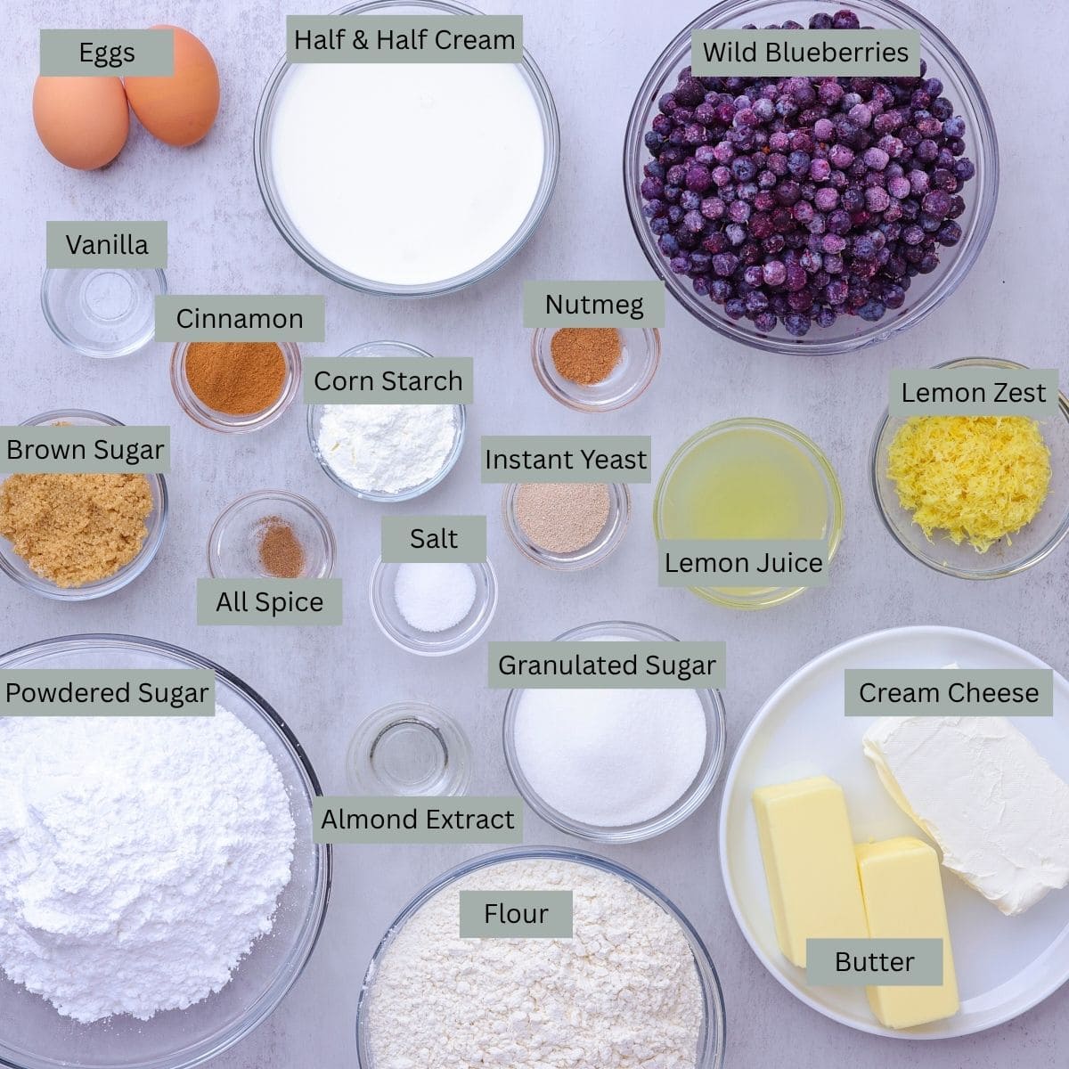 A visual display of the ingredients needed for the Blueberry Cinnamon Rolls - eggs, milk, blueberries, almond extract, cinnamon, corn starch, nutmeg, brown sugar, all spice, salt, quick rise yeast, lemon juice, lemon zest, cream cheese, butter, flour, sugar, vanilla and powdered sugar - each in their own bowl.