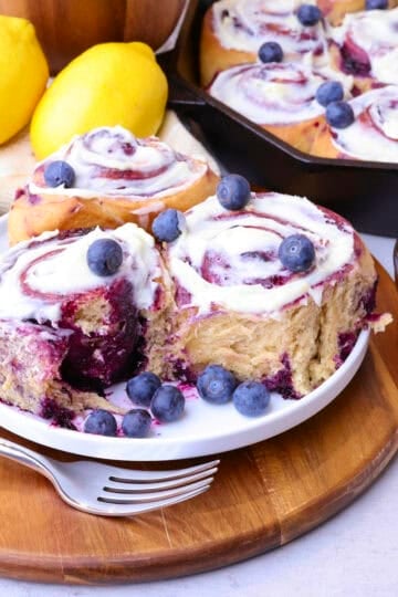 A trio of Blueberry Cinnamon Rolls on a plate with one bitten into. A filled skillet behind and lemons, blueberries, a fork and sauce arranged around them.