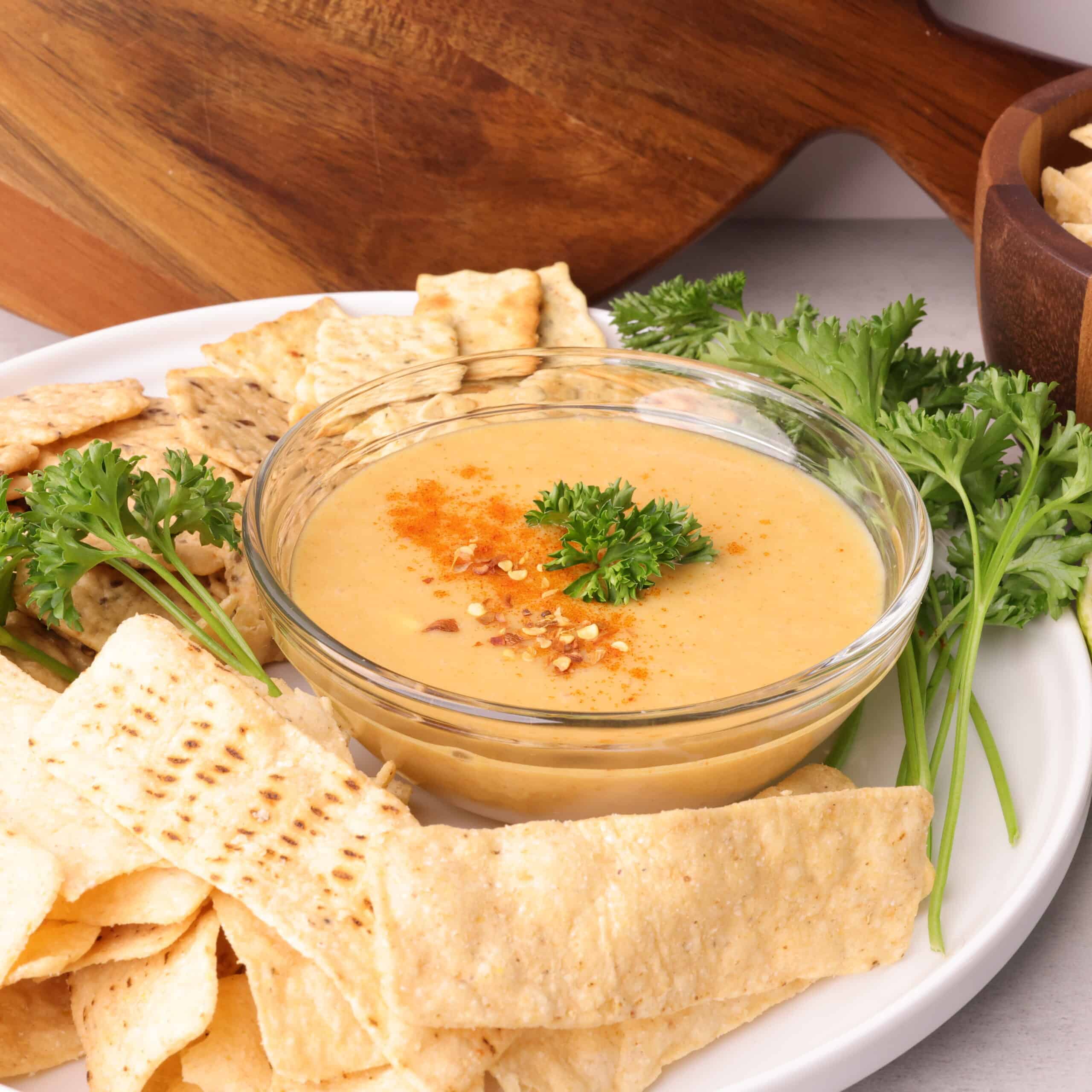 A bowl of the Honey Mayo Mustard Sauce on a plate with chips, crackers and greens.