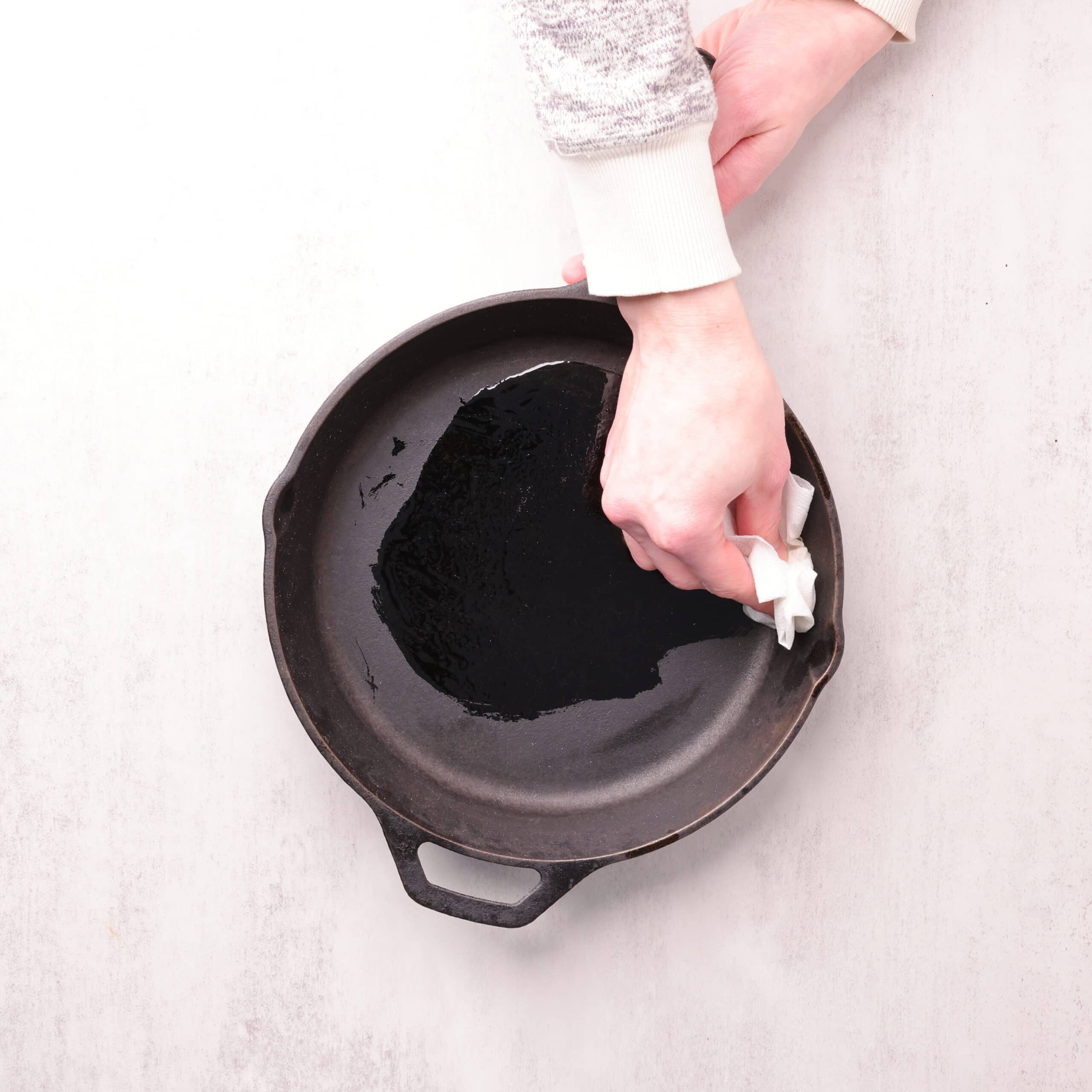 A skillet with oil in it and a hand spreading it around with paper towel.