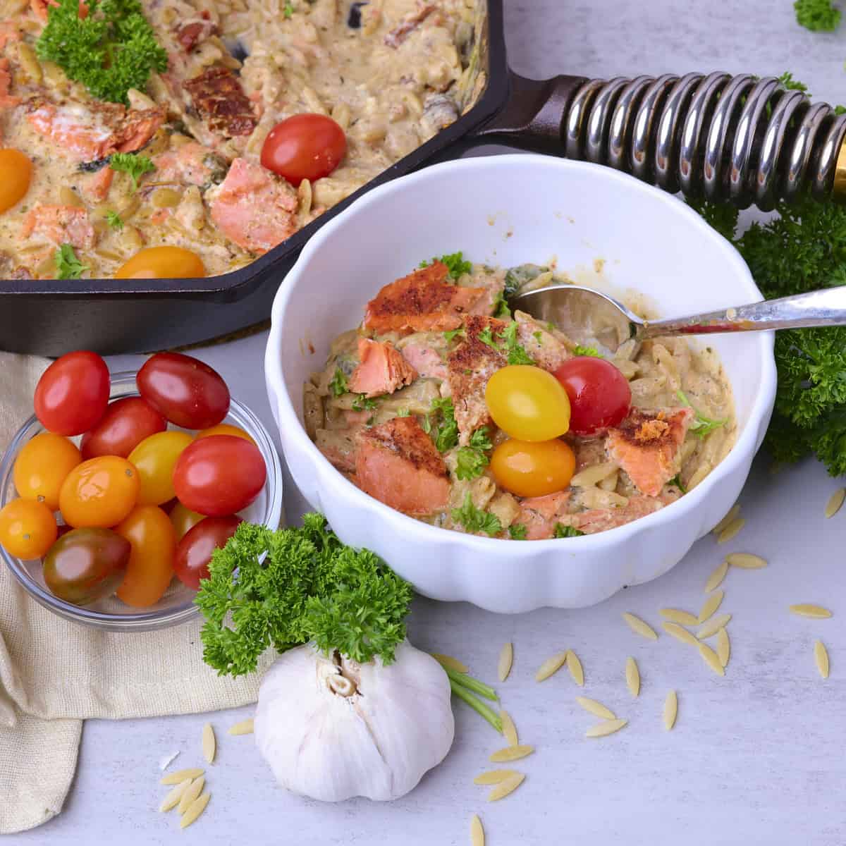 A bowl of the Salmon Pasta displayed by the skillet of pasta with a bowl of tomatoes, greens and head of garlic beside.