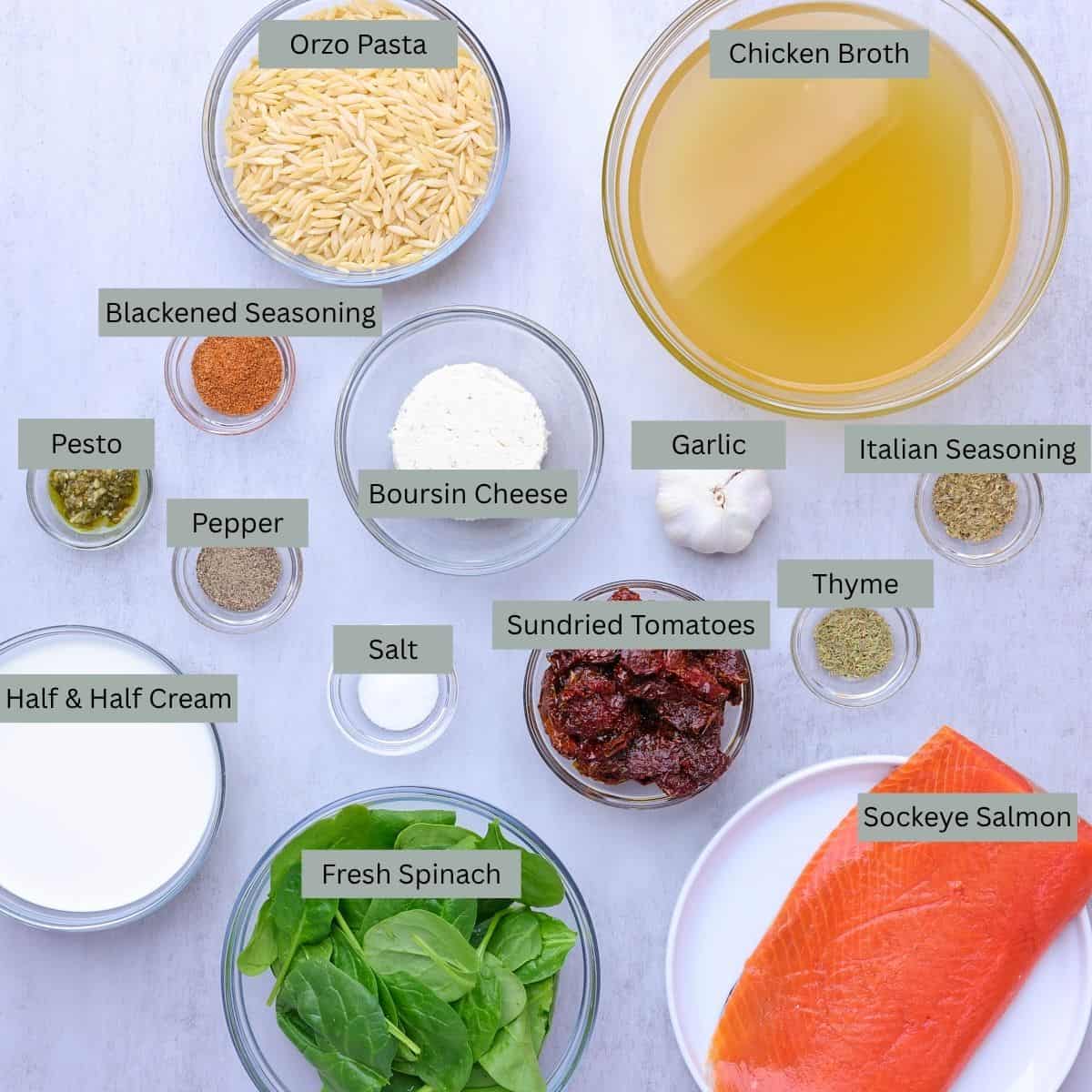 A visual display of the ingredients needed for the Salmon Pasta - orzo, chicken broth, blackened seasoning, pesto, pepper, salt, Boursin Cheese, sundried tomatoes, garlic head, thyme, Italian seasoning, half & half cream, spinach, salmon - each displayed in their own bowl.