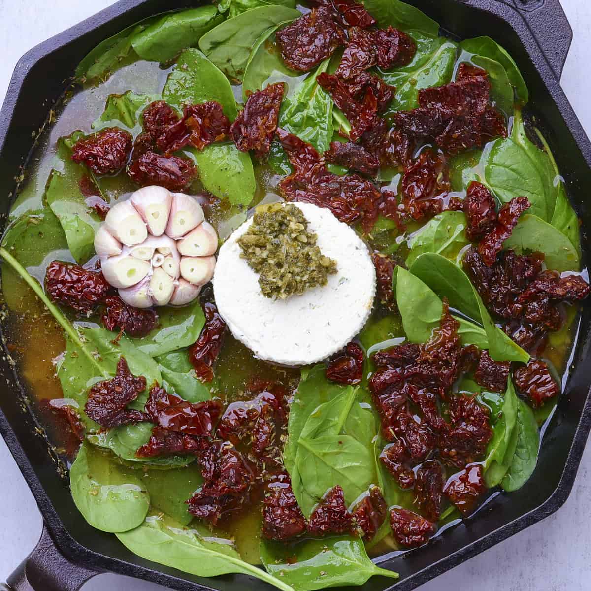A skillet with the Boursin cheese topped with pesto and garlic head in the middle with the fresh spinach and sundried tomatoes around it with the remaining ingredients underneath.