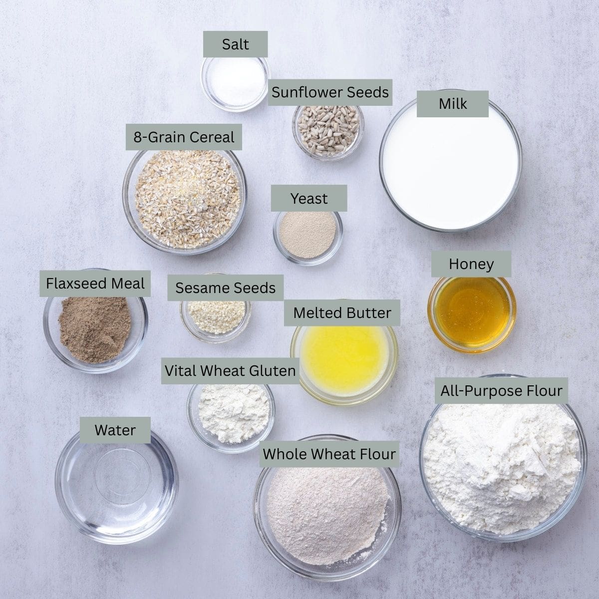 A visual display of the ingredients needed to bake the Seeded Bread - milk, honey, all purpose flour, whole wheat flour, melted butter, sesame seeds, sunflower seeds, salt, 7-grain cereal, yeast, vital wheat gluten, water, flaxseed meal - each in their own bowl.