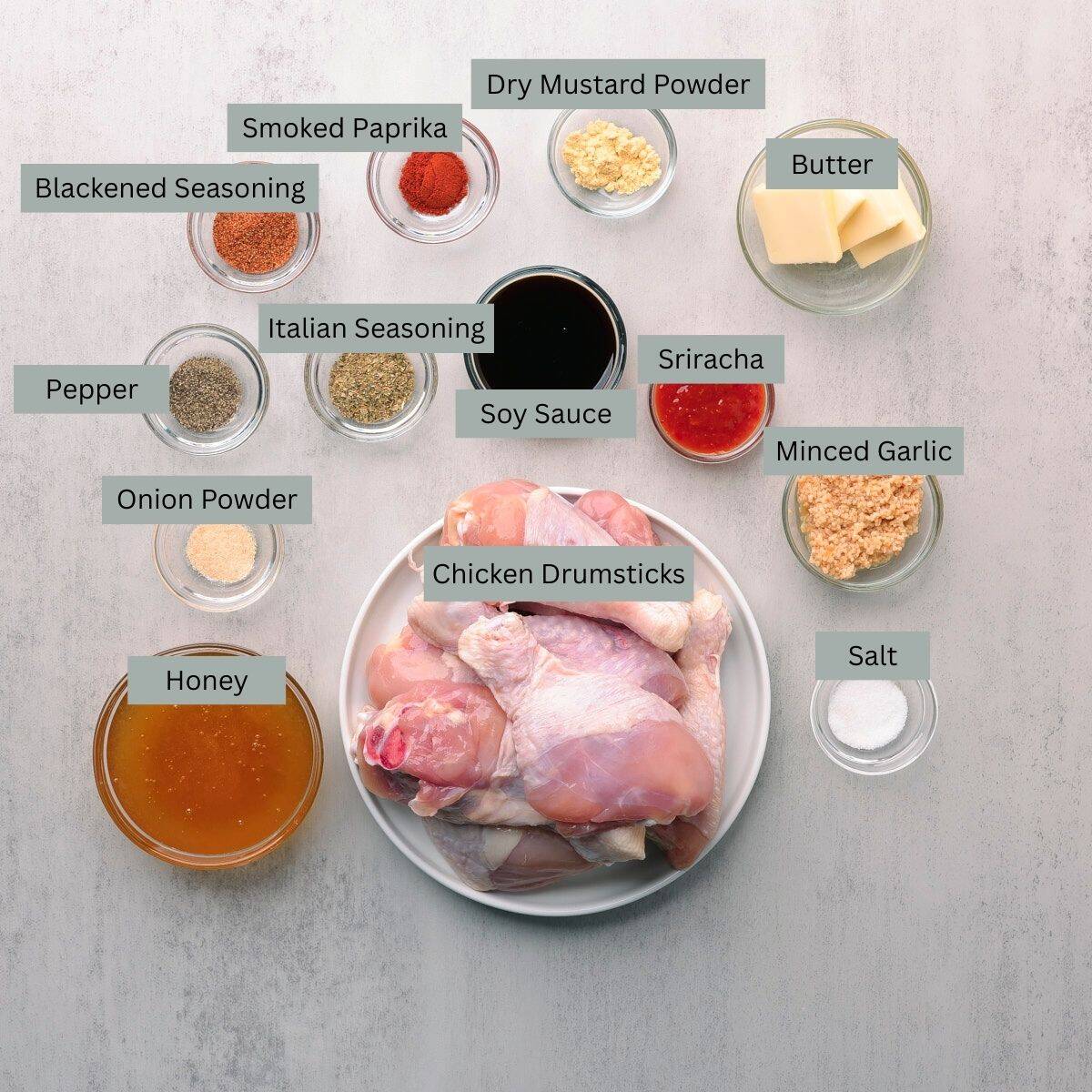 A visual display of the ingredients needed for the Sticky Chicken Drumsticks - drumsticks, honey, minced garlic, sriracha, soy sauce, salt, pepper, onion powder, Italian seasoning, smoked paprika, blackened seasoning, dry mustard and butter - each in their own bowl.