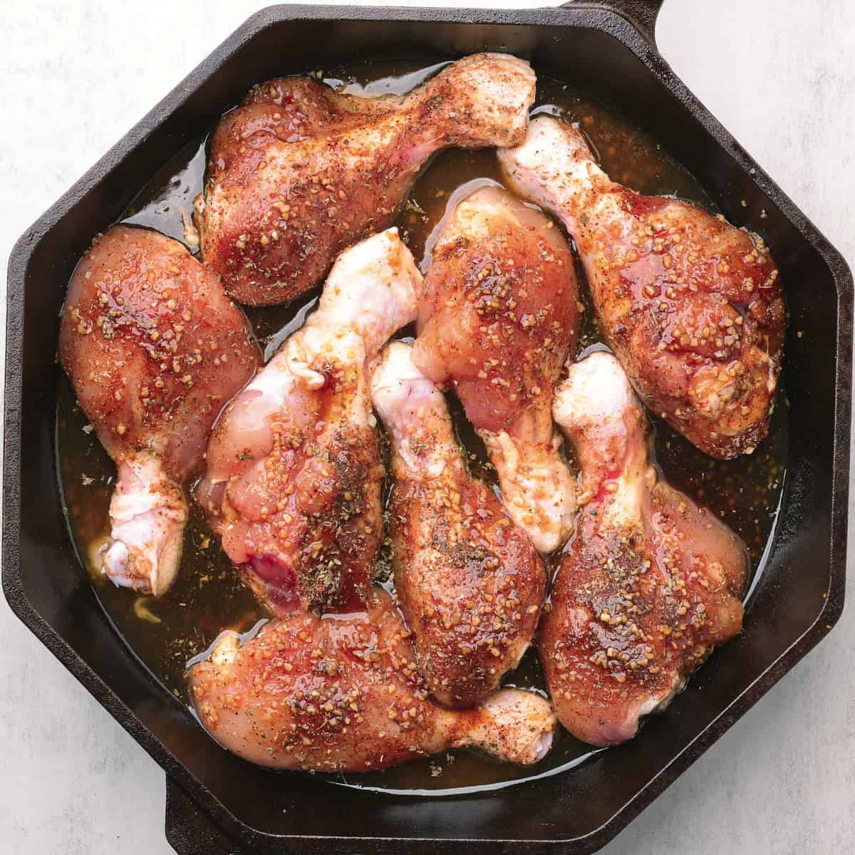 A skillet with the seasoned, raw drumsticks arranged in the melted sauce, ready to bake.