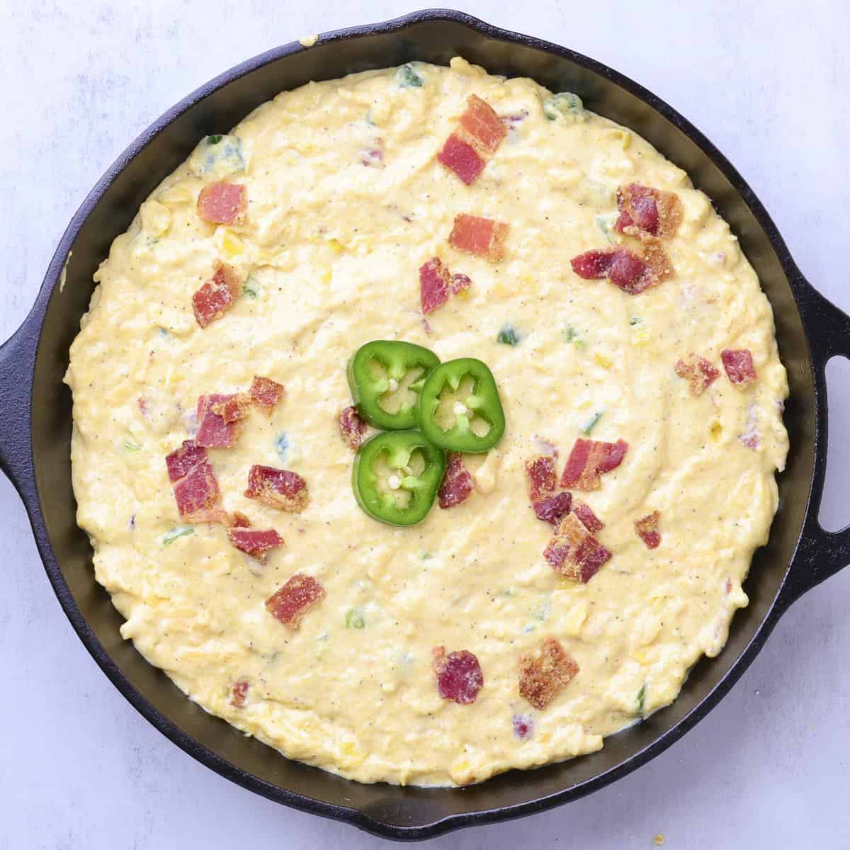 A skillet with the Cheddar Cheese Jalapeno Cornbread batter in it topped with jalapeno slices and bacon bits.