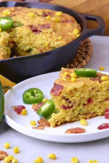 A slice of Cheddar Cheese Jalapeno Cornbread on a plate with a skillet of the cornbread in the background.