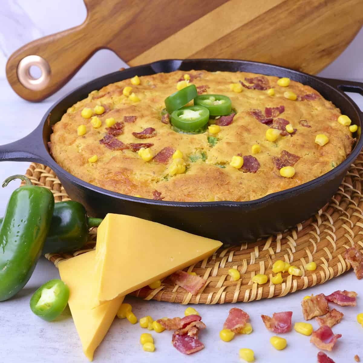 The fully baked Cheddar Cheese Jalapeno Cornbread in a skillet and displayed with jalapenos, cheddar cheese and bacon bits.