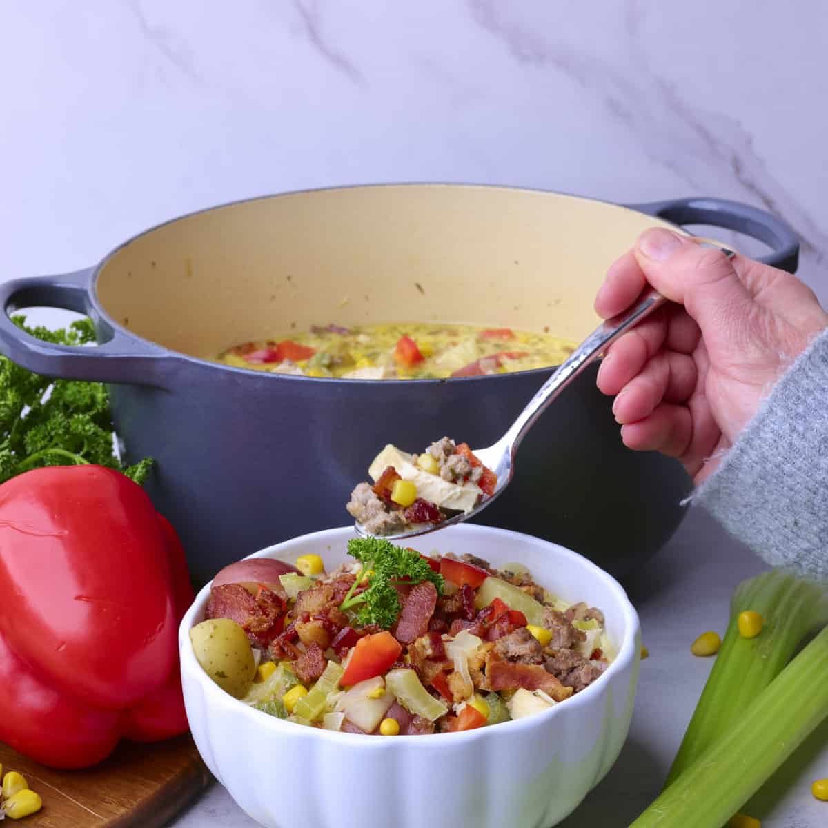 A bowl of Chicken and Sausage Soup with a hand holding a spoonful above it with the pot of soup behind and vegetables around.
