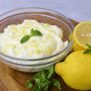 A bowl of Lemon Cream Cheese Icing topped with zest and mint leaves placed displayed with lemons and mint beside.