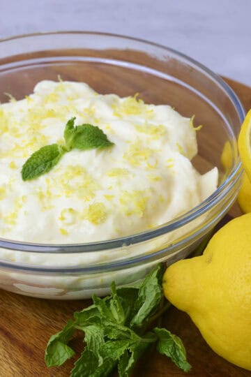 A bowl of Lemon Cream Cheese Icing topped with zest and mint leaves placed displayed with lemons and mint beside.
