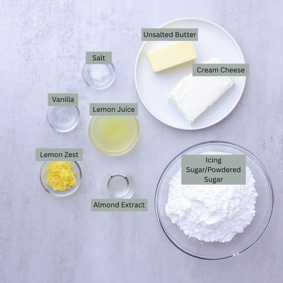 A visual display of the ingredients needed for the Lemon Cream Cheese Icing - butter, cream cheese, icing sugar (powdered sugar), salt, vanilla, almond extract, lemon juice and lemon zest - each displayed in their own bowl.
