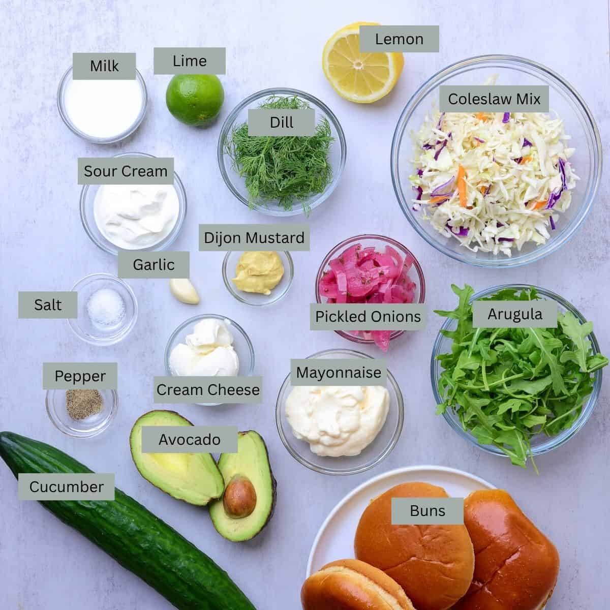 A visual display of the ingredients needed for the Salmon Burgers made with Canned Salmon - milk, lime, lemon, dill, coleslaw mix, arugula, pickled onions, Dijon mustard, sour cream, salt, pepper, clove of garlic, cream cheese, mayonnaise, avocado, cucumber and buns.