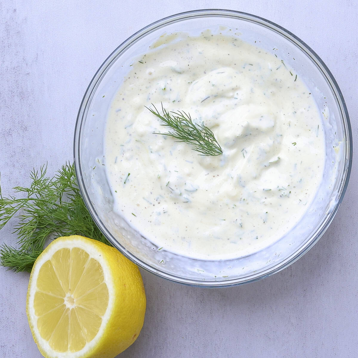 A bowl of lemon-dill aioli displayed with a sprig of dill and half a lemon.